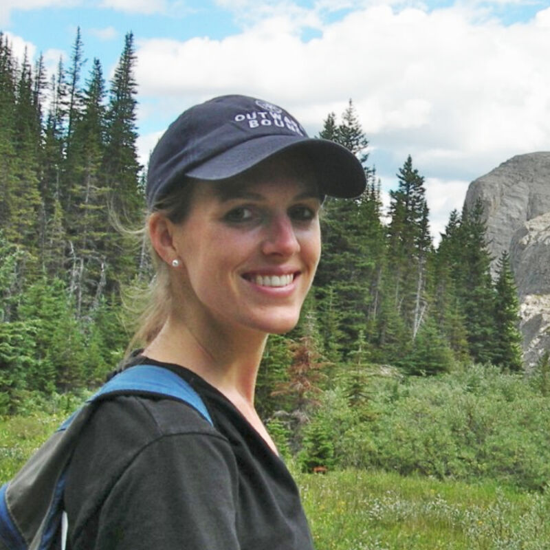 The image shows a woman wearing a cap and a backpack, smiling at the camera. She is in a natural environment, with trees and a mountain in the background. The lighting suggests it's daytime, and the overall impression is of a hike or outdoor activity.